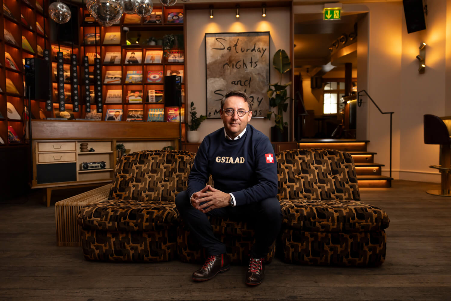 Michel Wichman auf einem gemusterten Sofa in einem Raum mit Bücherregalen und warmen Wandlampen.