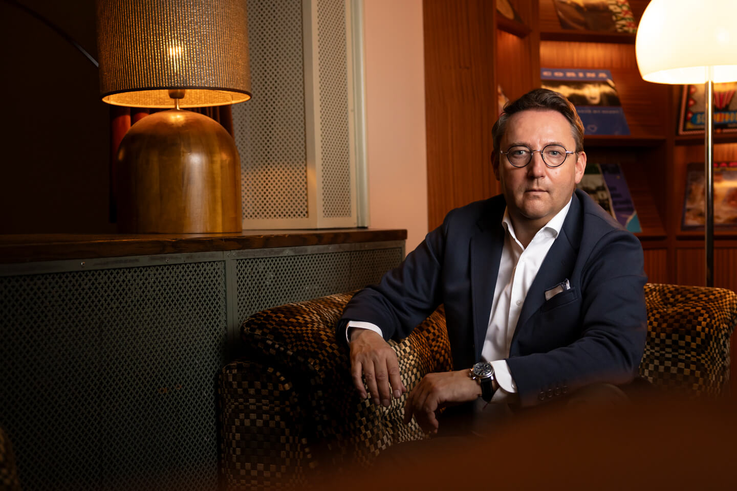 Michel Wichman in dunklem Anzug mit weißem Hemd sitzt auf einem gemusterten Sofa in einem Raum mit warmem Licht.