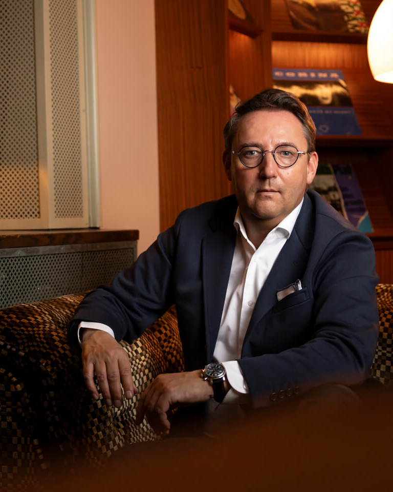 Michel Wichman in dunklem Anzug mit weißem Hemd sitzt auf einem gemusterten Sofa in einem Raum mit warmem Licht.