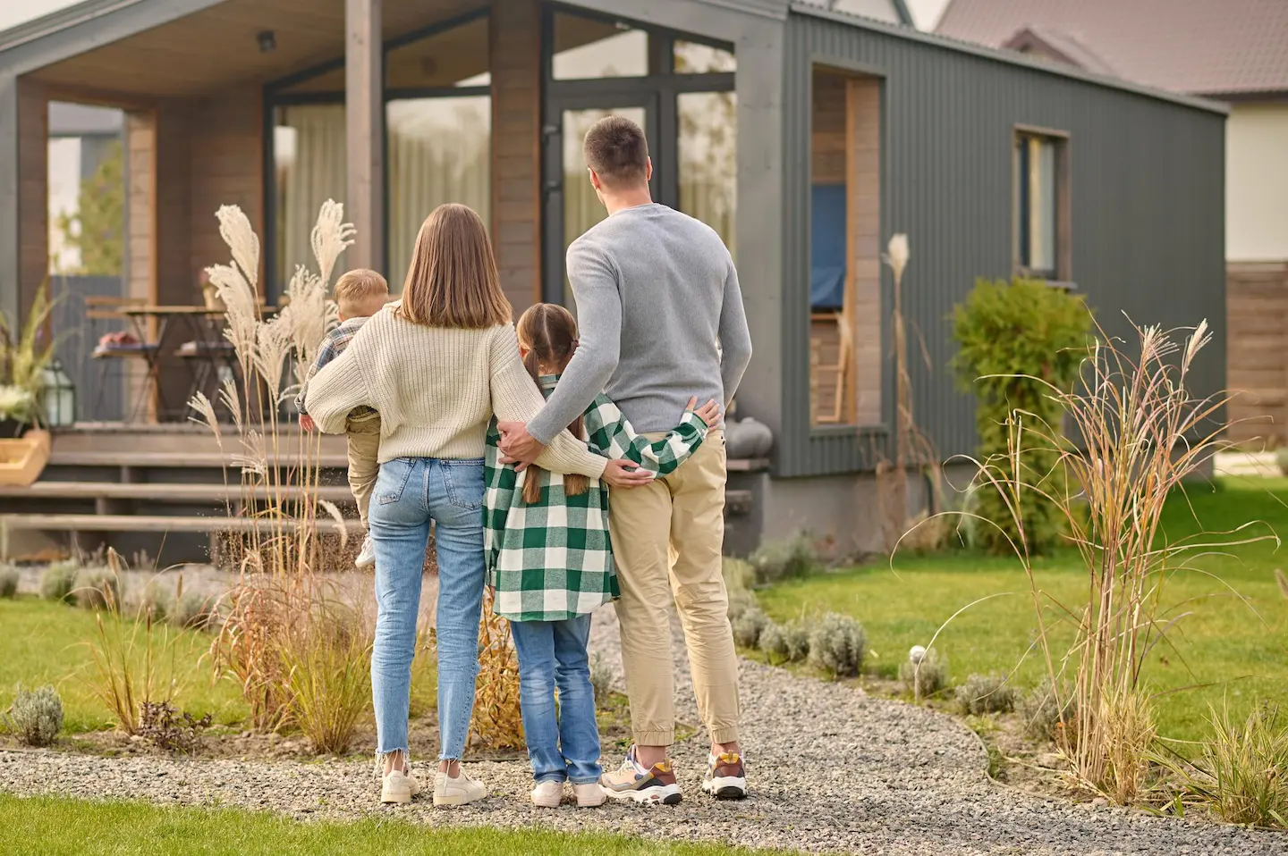 Une famille de quatre personnes vue de dos, marchant vers une maison moderne avec un jardin.
