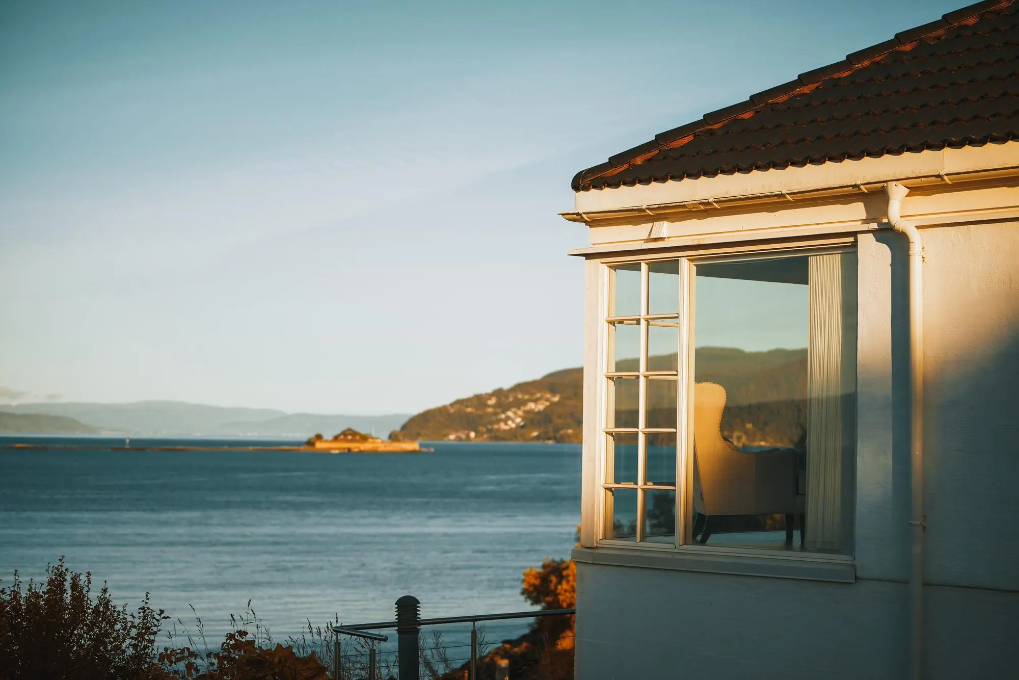 Fauteuil visible à travers une grande fenêtre d'une maison surplombant une étendue d'eau avec une île et des collines au loin au coucher du soleil.