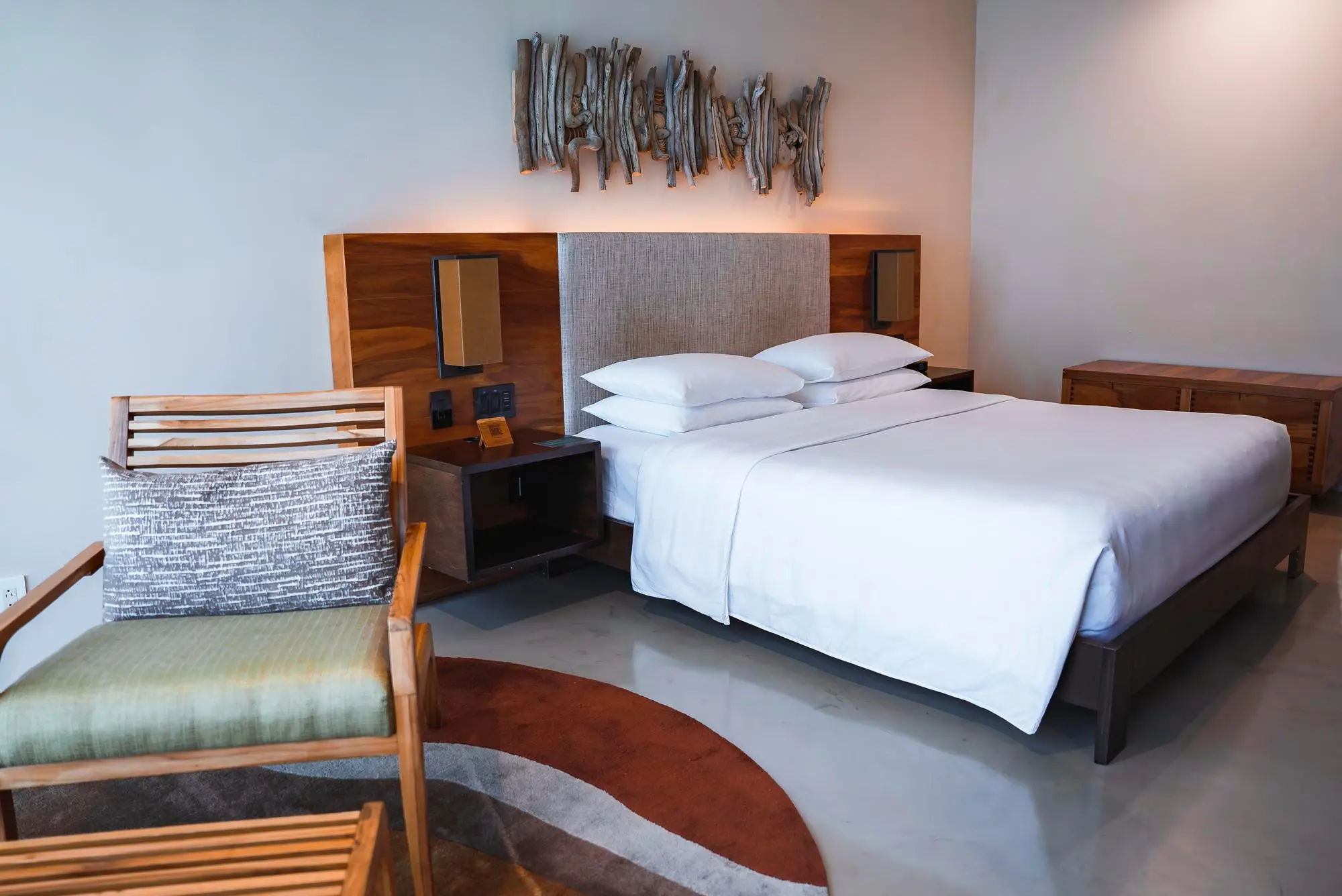 Chambre avec lit double aux draps blancs, table de chevet en bois, fauteuil en bois avec coussin et tapis rond coloré.