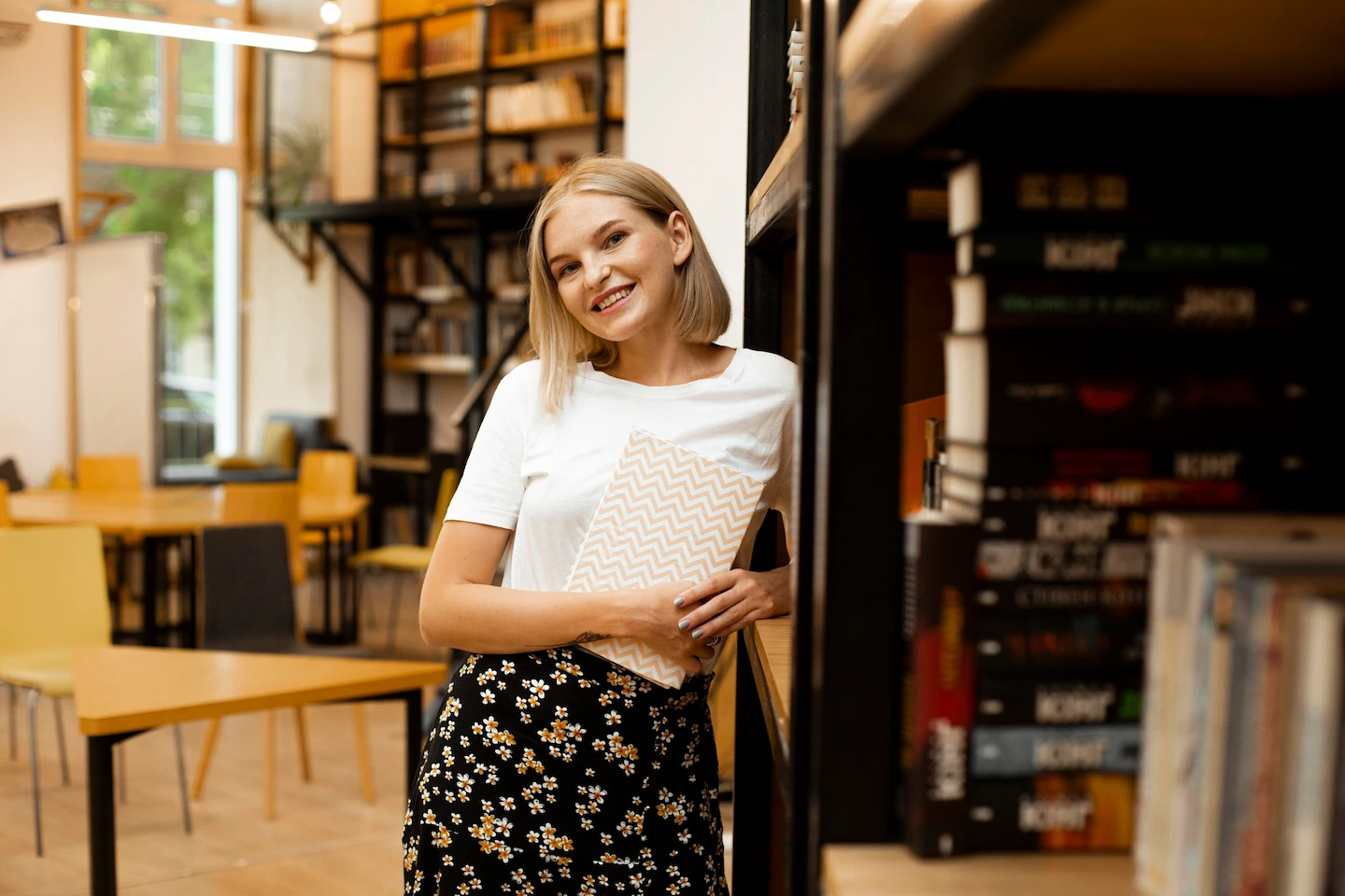 Jeune femme souriante tenant un cahier debout près d'une étagère dans une bibliothèque lumineuse.