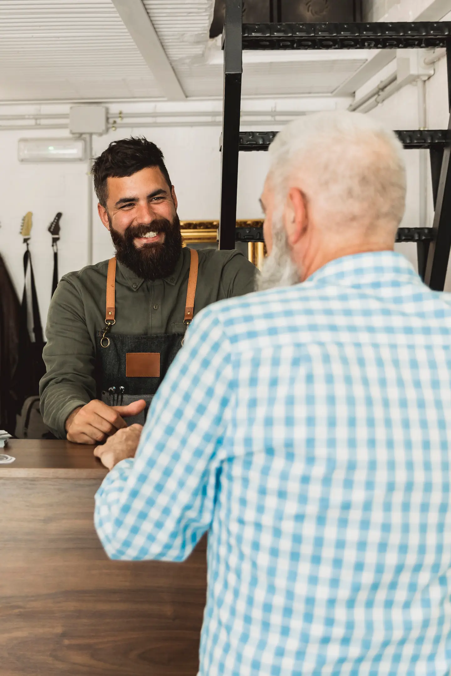 Un barbier souriant portant un tablier serrant la main d'un homme âgé en chemise à carreaux bleue.