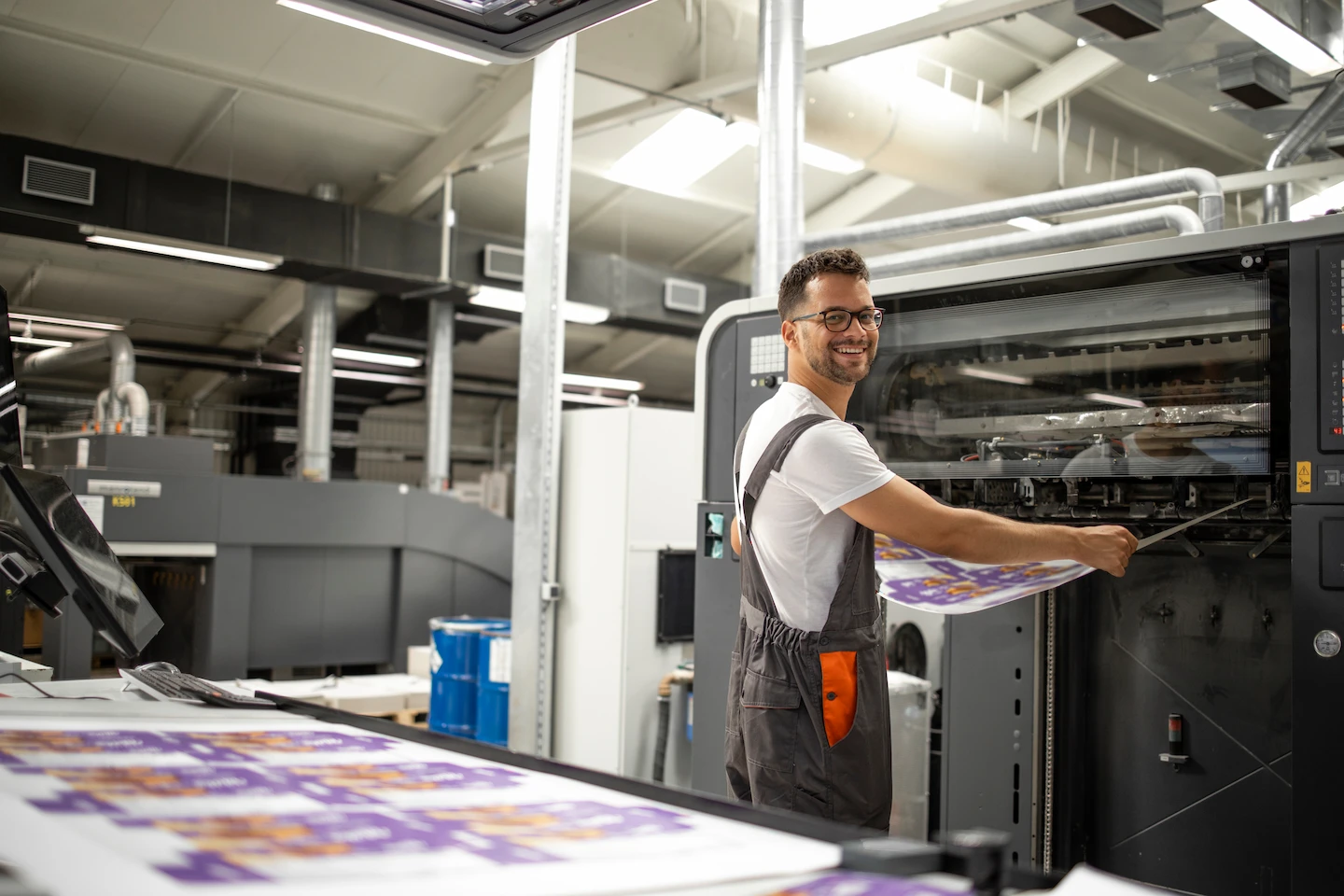 Un homme souriant en salopette tient une grande feuille imprimée devant une machine d'impression industrielle dans une usine.