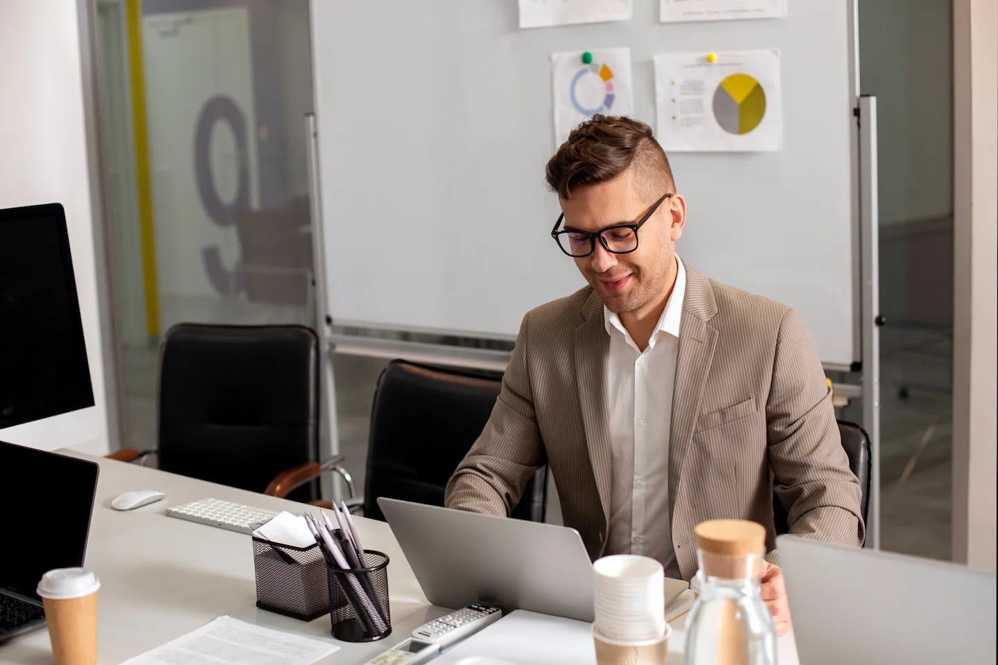 Homme en costume rayé beige travaillant sur un ordinateur portable dans un bureau moderne avec tableau blanc et accessoires de bureau.