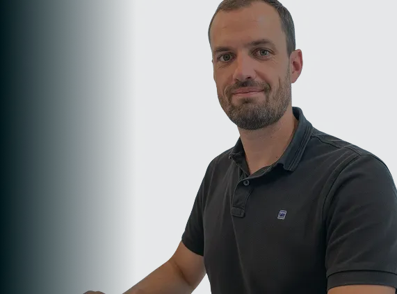 Homme souriant portant un polo noir sur fond sombre.