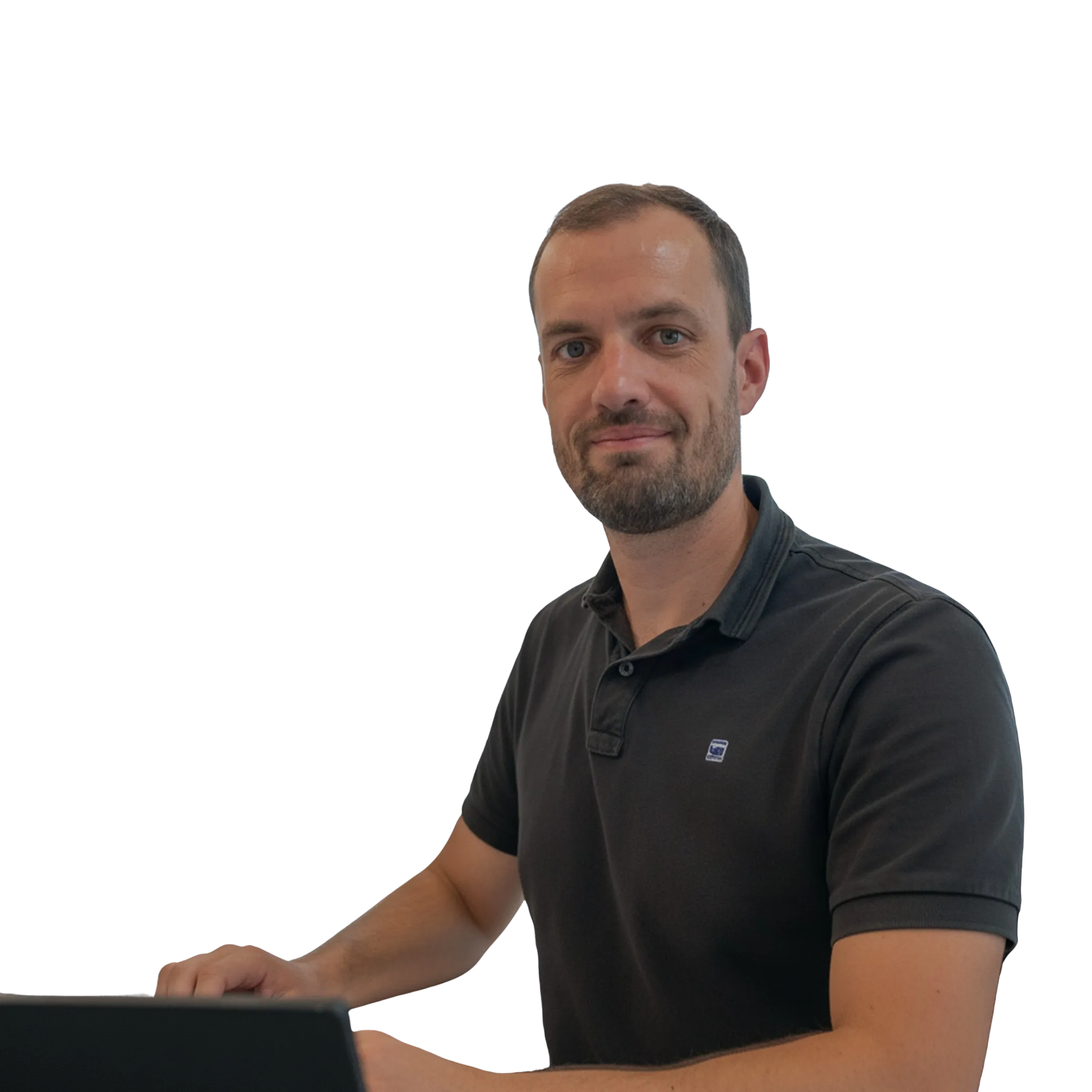 Homme souriant portant un polo noir, assis devant un ordinateur portable.