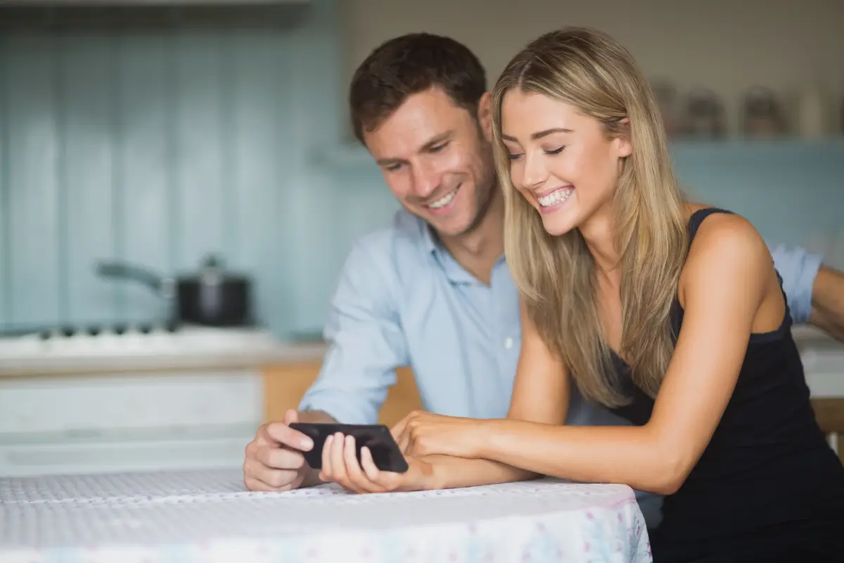 Un homme et une femme souriants regardant ensemble un téléphone portable à une table couverte d'une nappe.