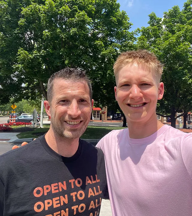 Two men smiling outdoors with green trees and a street in the background, one wearing a black shirt with 'OPEN TO ALL' text.