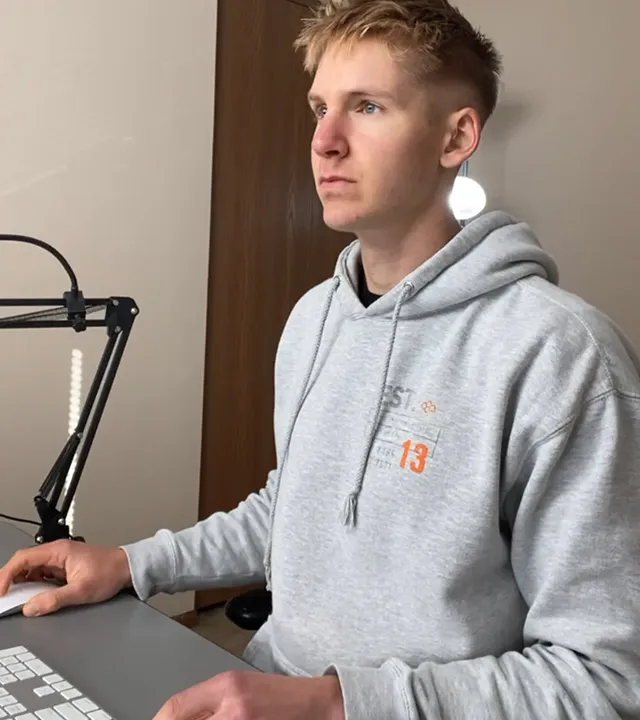 Young man in a gray hoodie sitting at a desk, using a keyboard with a focused expression.