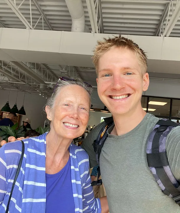 Smiling elderly woman and young man taking a selfie together indoors, the woman wearing a blue striped cardigan and the man with a backpack.