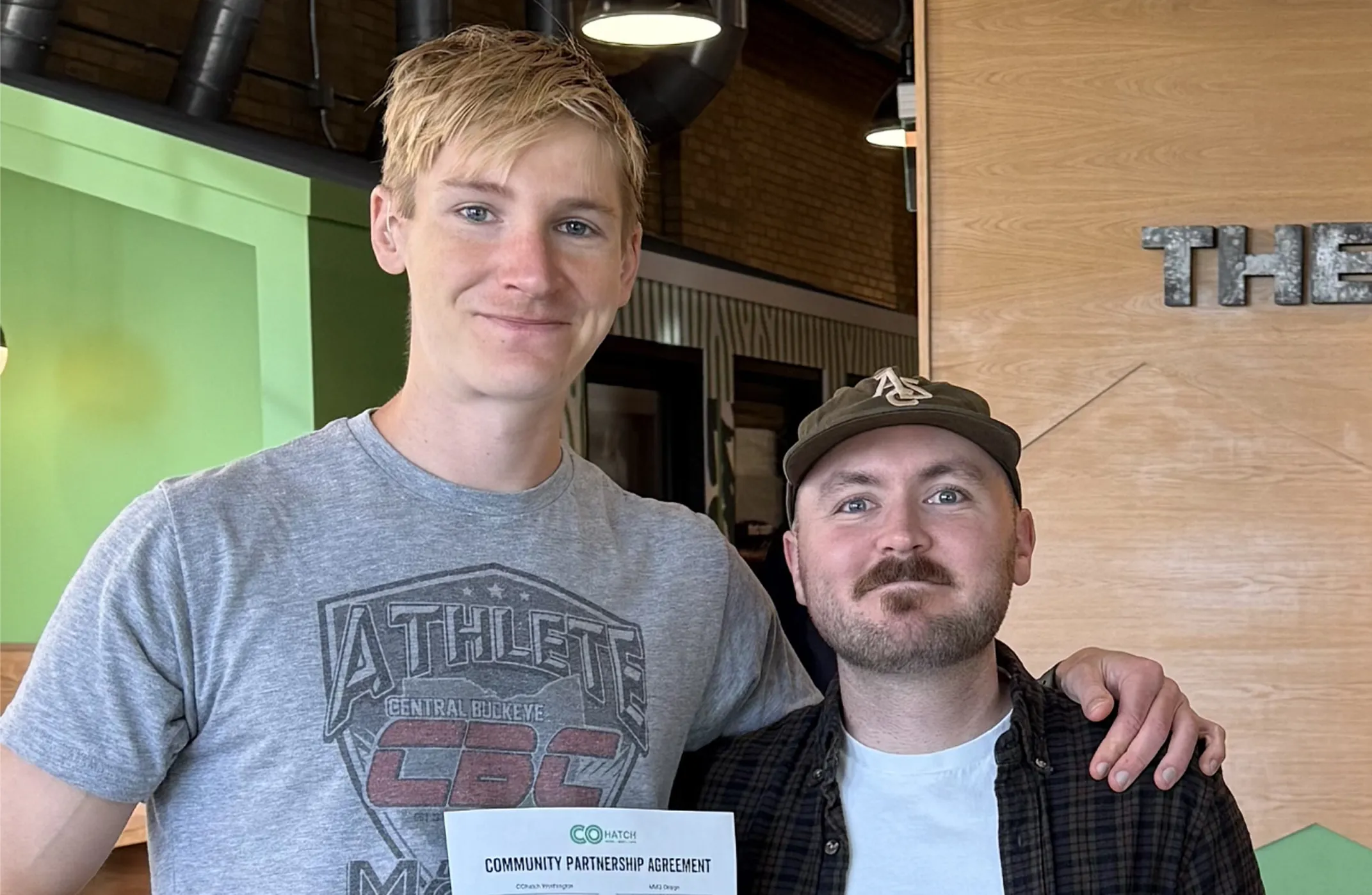Two men standing together indoors, one with an arm around the other, holding a community partnership agreement document.