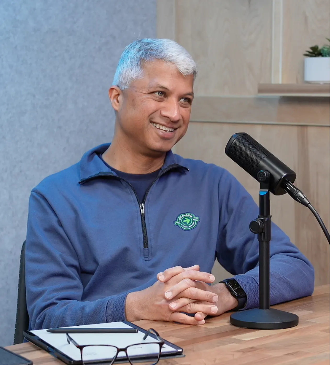 Smiling man with gray hair sitting at a table with a microphone, clipboard, glasses, and pen.