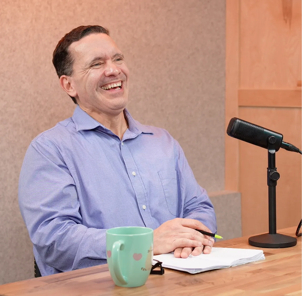 Man in a blue shirt laughing while sitting at a table with a microphone, notes, and a mug.