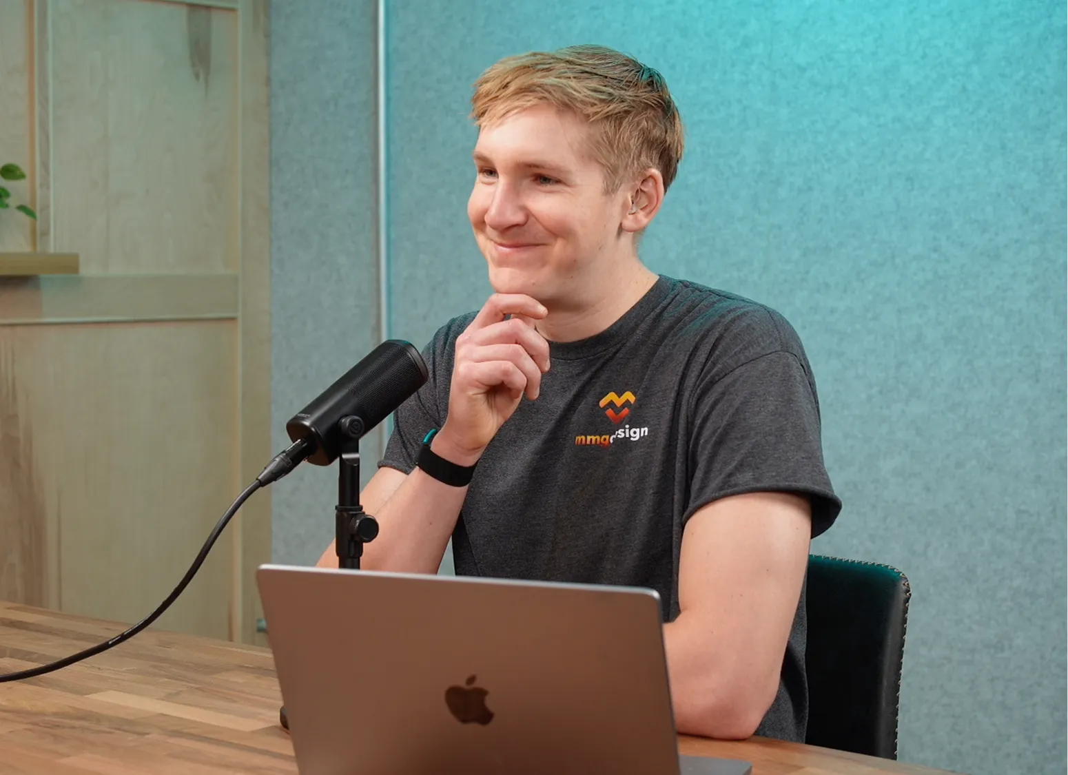 Man sitting at wooden table with laptop and microphone, resting chin on hand, smiling thoughtfully.