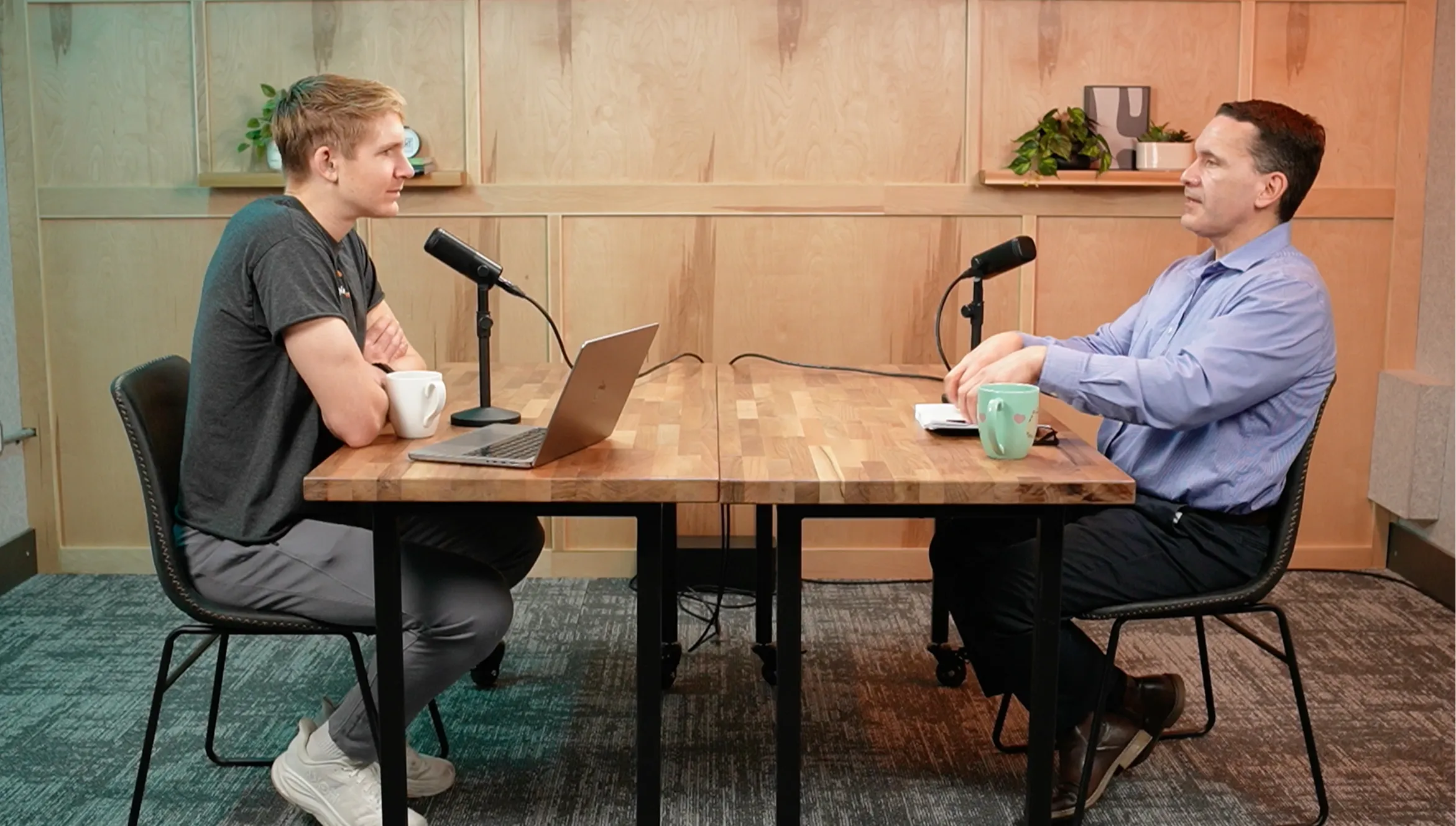 Two men seated across a table with microphones, a laptop, mugs, and notes in a recording setup.