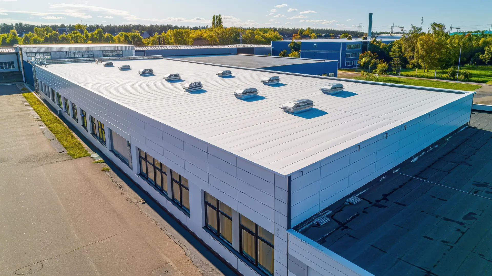 A large modern industrial or commercial building with a flat white roof featuring multiple ventilation domes, surrounded by greenery and blue skies.