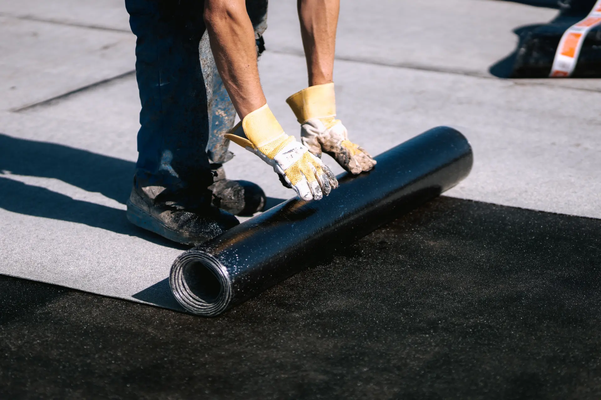 Worker wearing gloves and boots unrolling black roofing material on a flat roof surface.