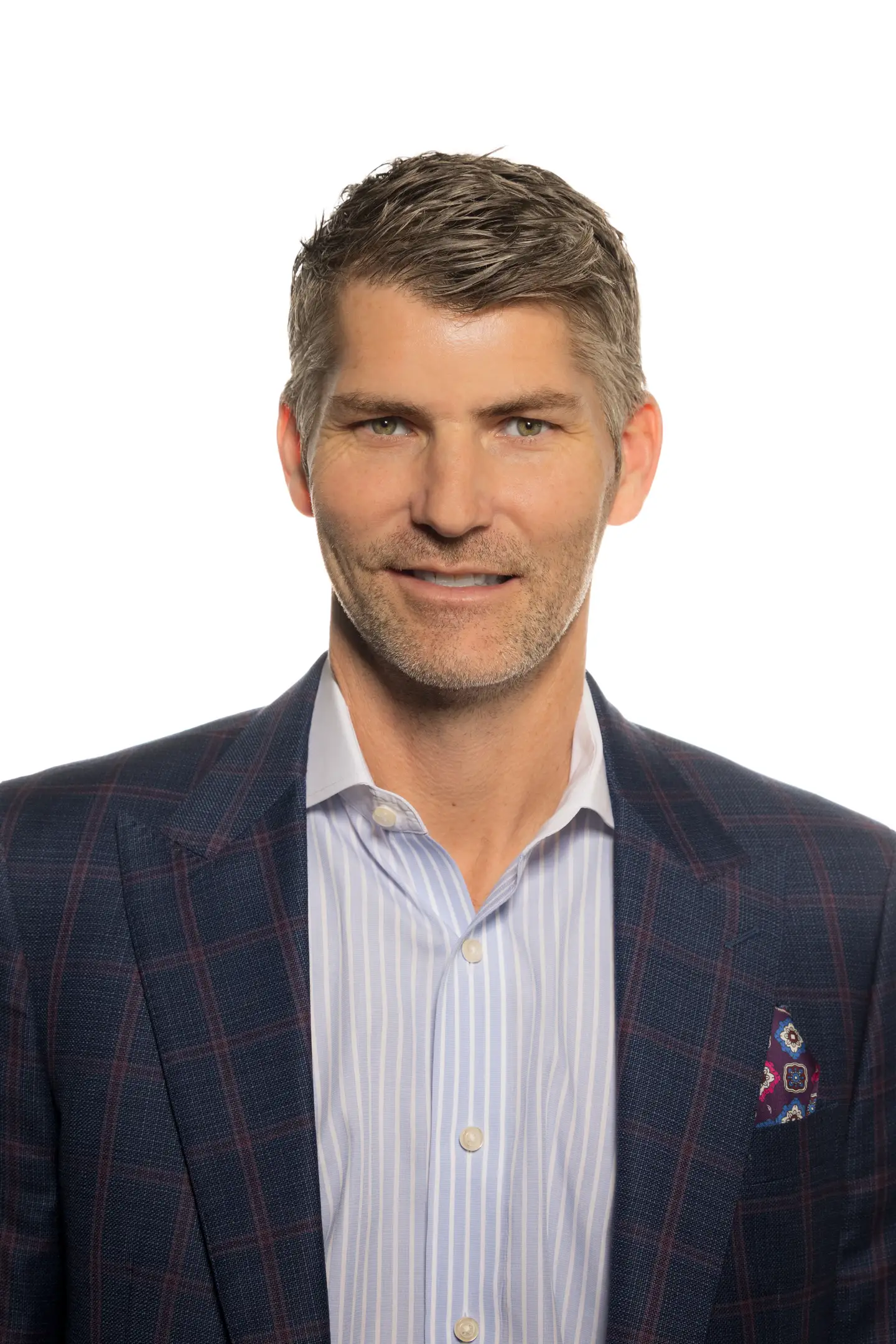 Middle-aged man with short gray hair wearing a blue plaid blazer, a light blue striped shirt, and a patterned pocket square, smiling slightly against a white background.