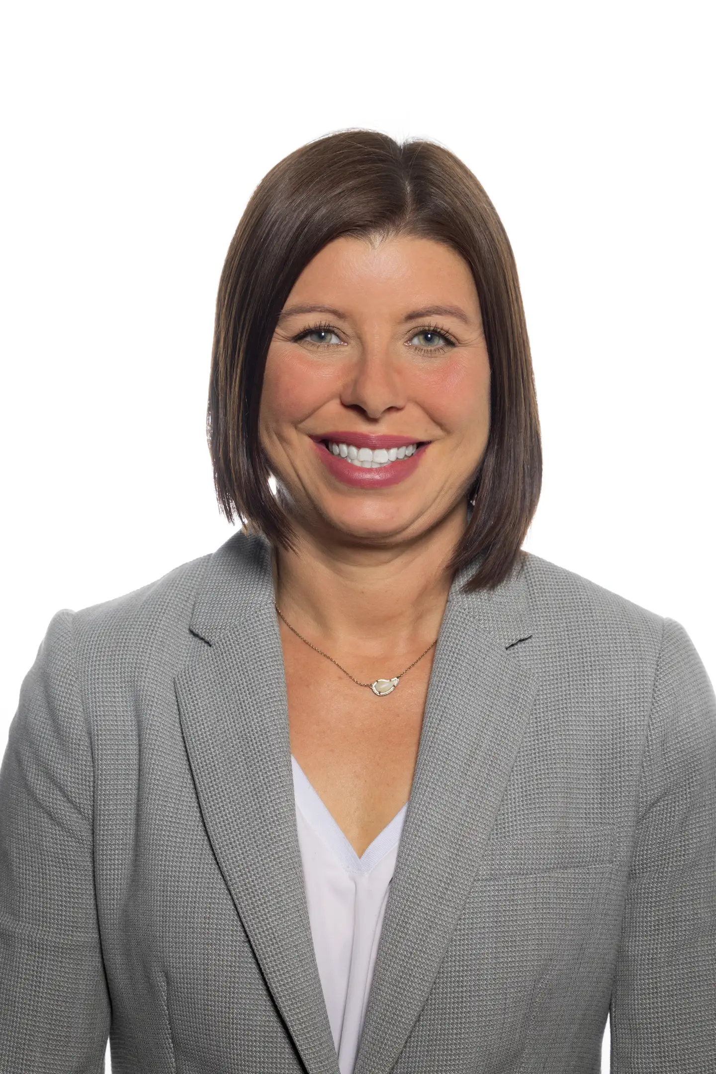 Smiling woman with straight brown hair wearing a gray blazer and white blouse against a white background.