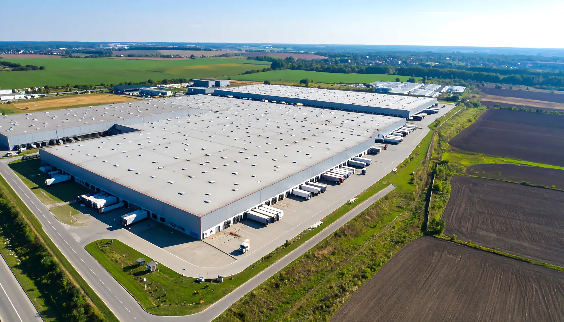 Aerial view of a large industrial warehouse complex surrounded by green fields and dirt plots.