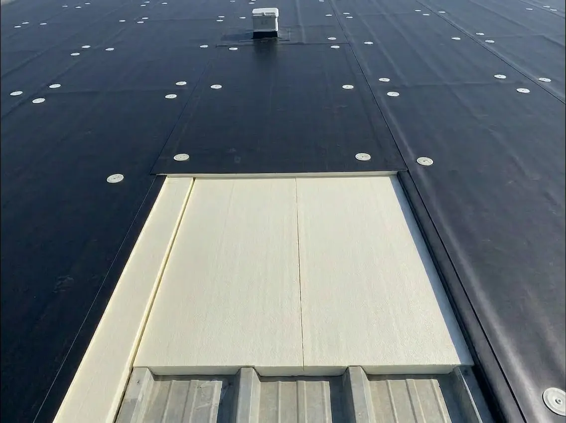 Black roofing membrane partially covering white insulation panels on a metal roof with a vent pipe in the background.