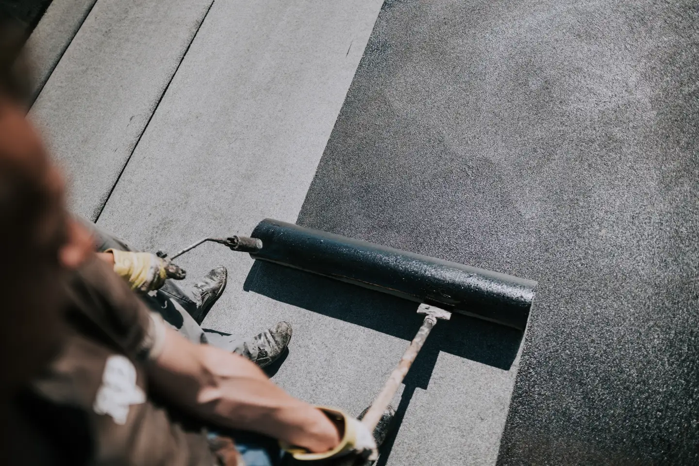 Worker using a large roller to apply black roofing material on a flat roof.