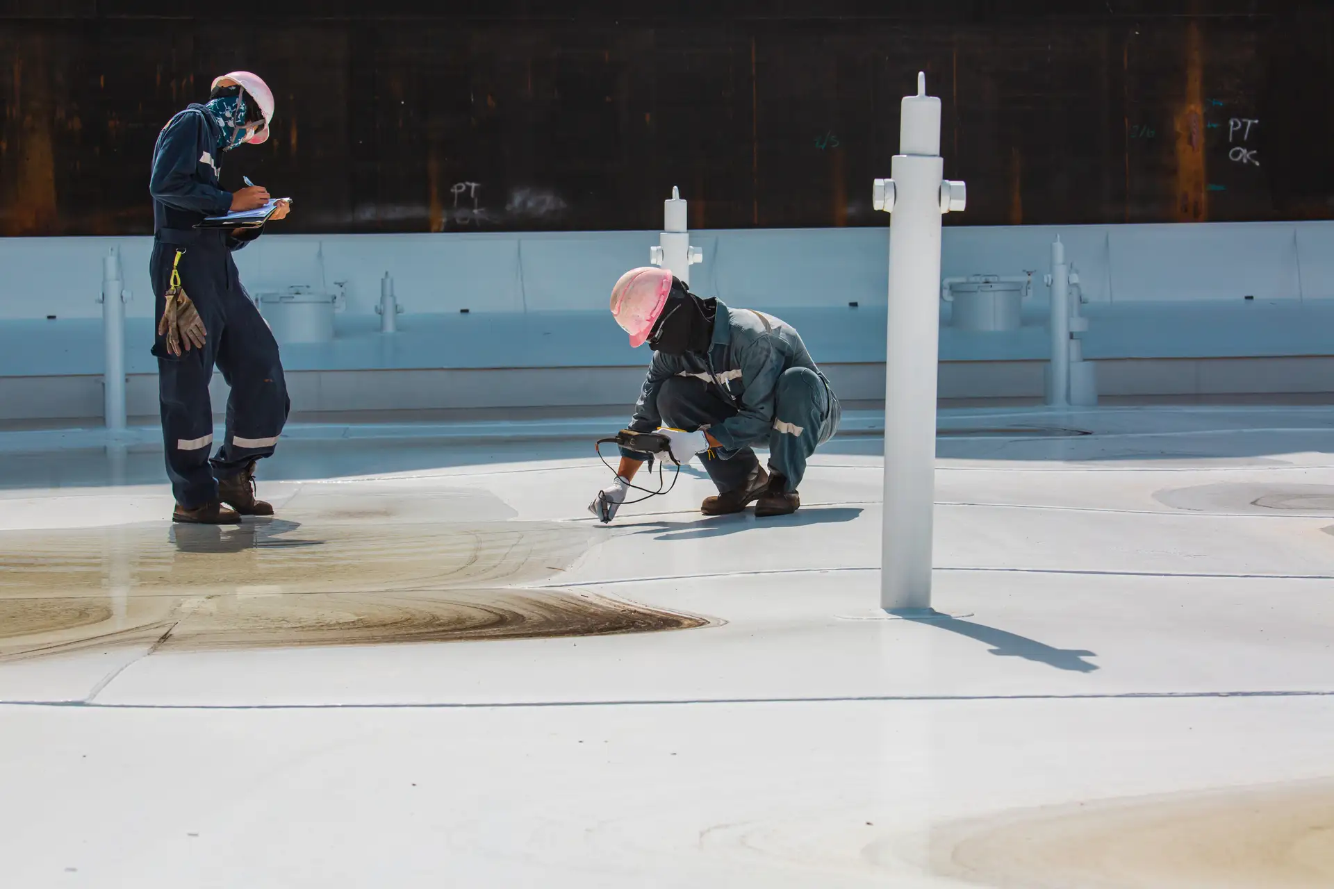 Two workers in protective gear inspecting and testing a white industrial tank surface, one writing on a clipboard and the other using a handheld device.