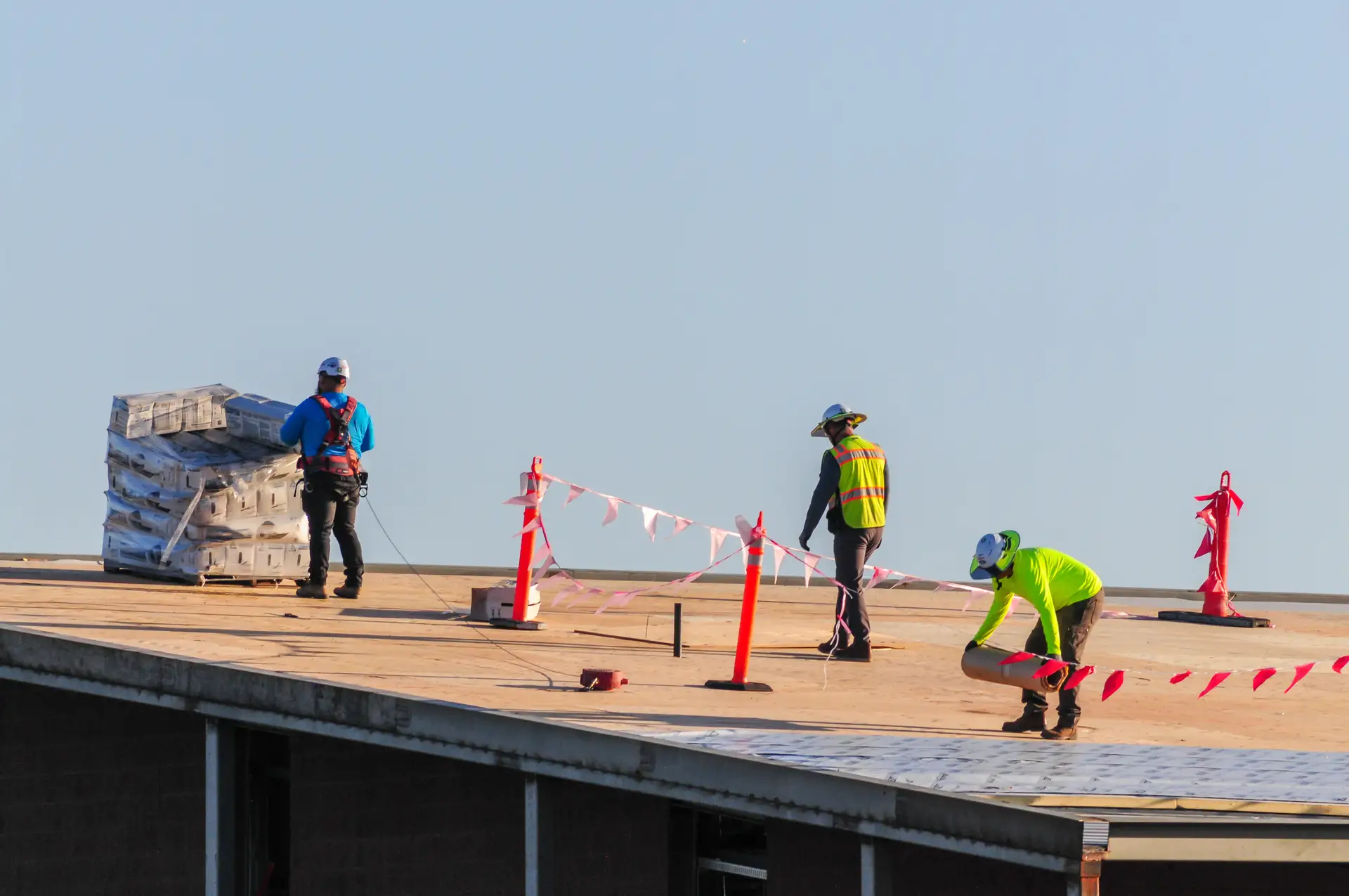 Three construction workers on a rooftop installing roofing materials with safety equipment and orange safety barriers.
