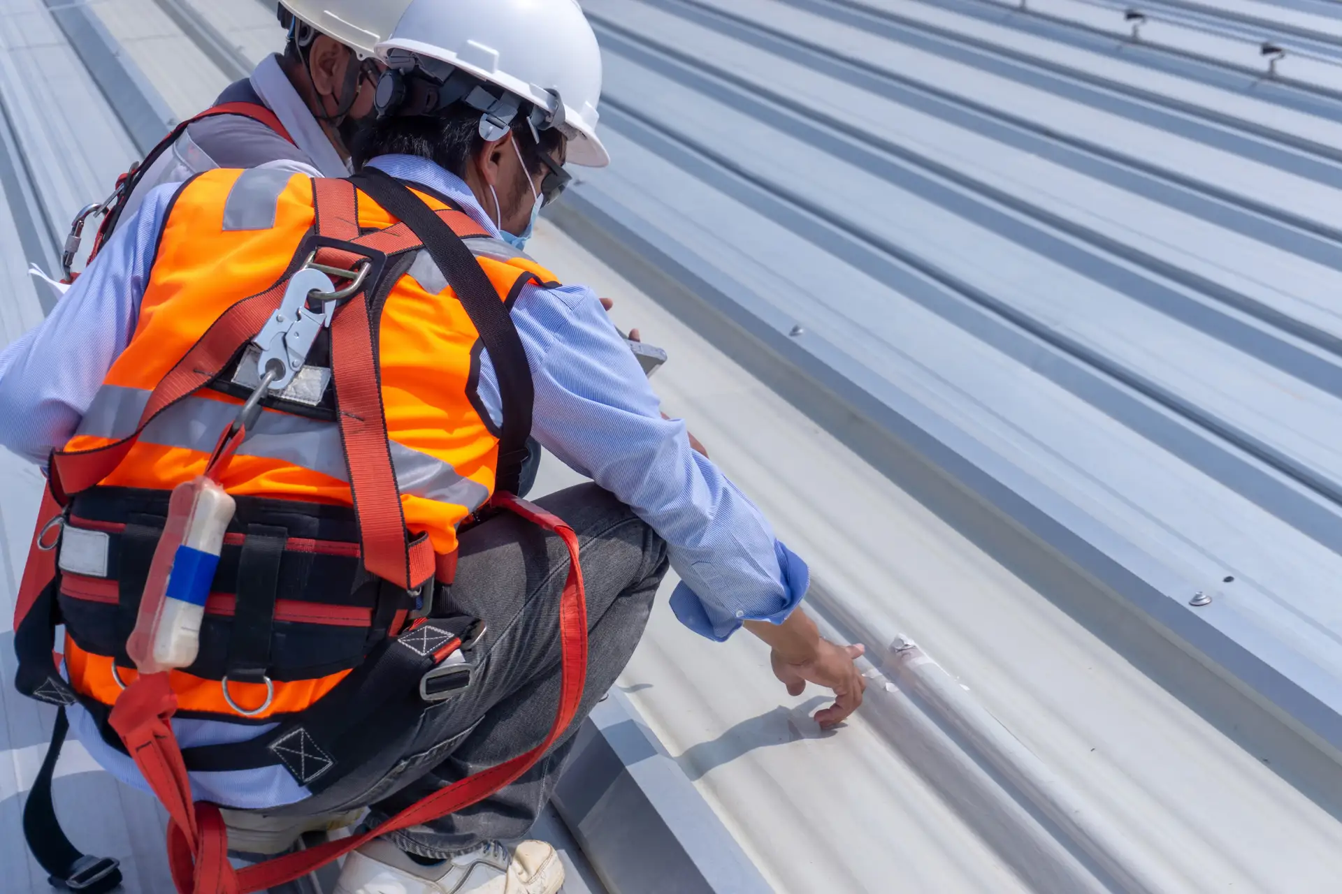 Two construction workers in safety helmets and orange vests inspecting a metal roof panel.