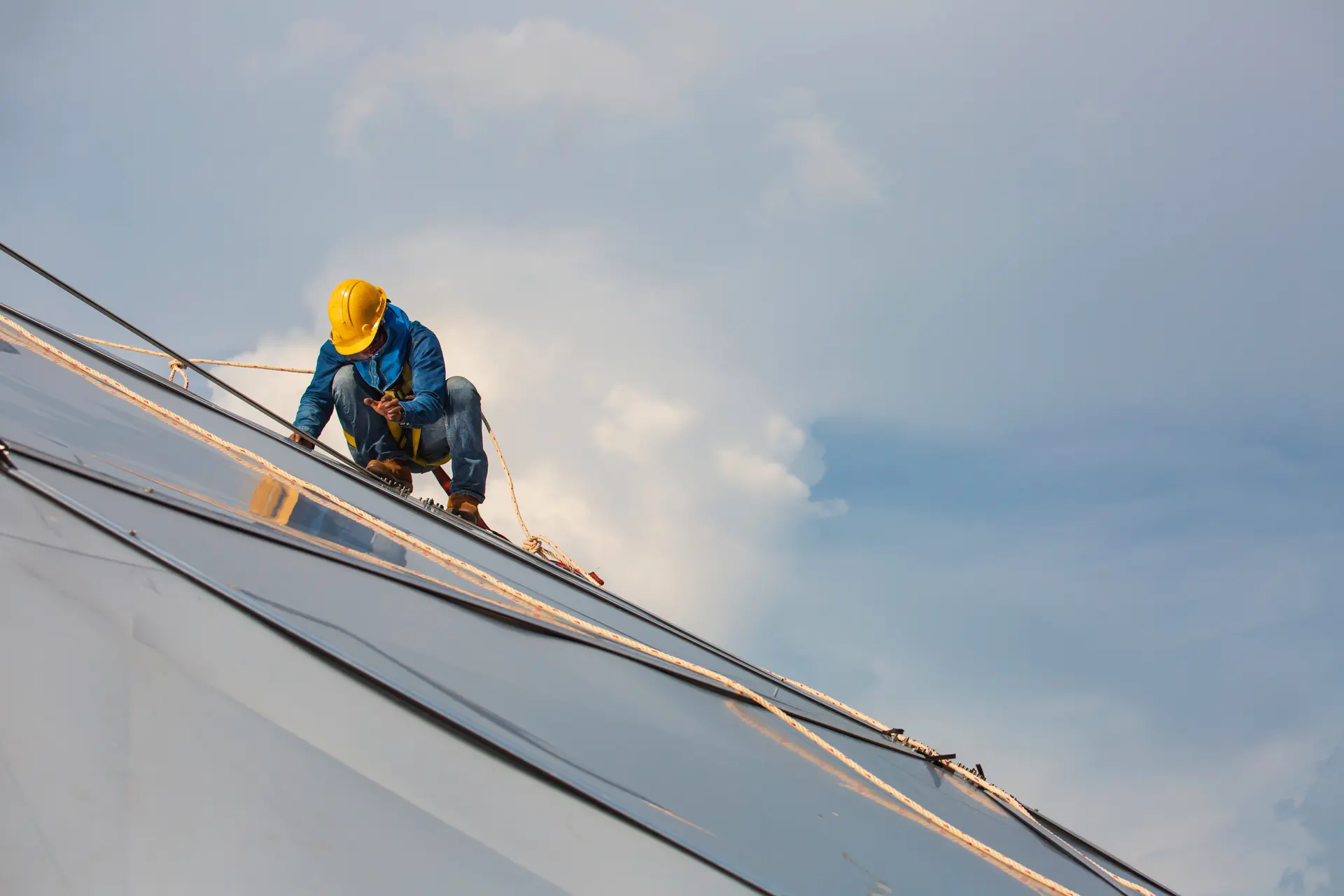 Worker in a yellow hard hat and safety harness installing solar panels on a rooftop under a partly cloudy sky.