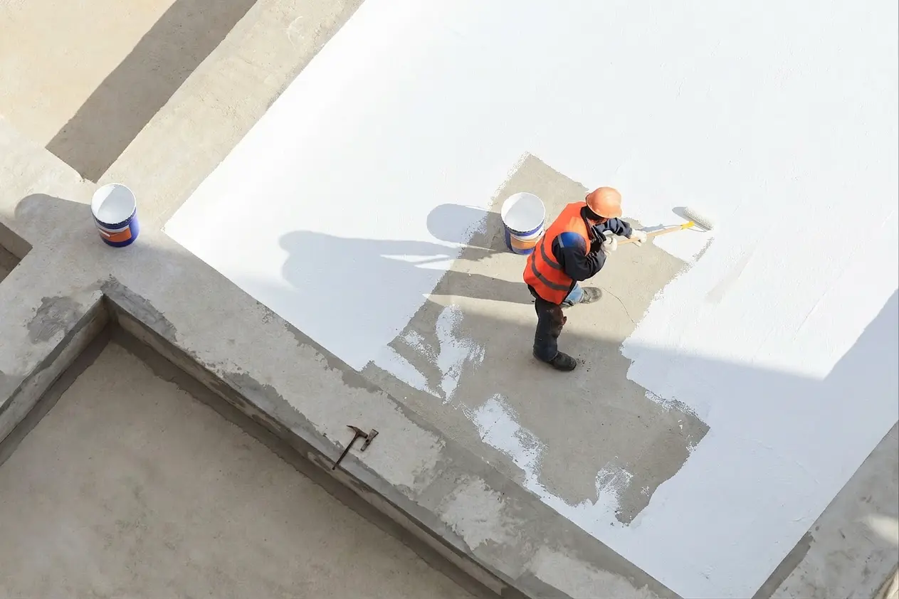 Construction worker in an orange vest and helmet painting a concrete surface white with a roller.