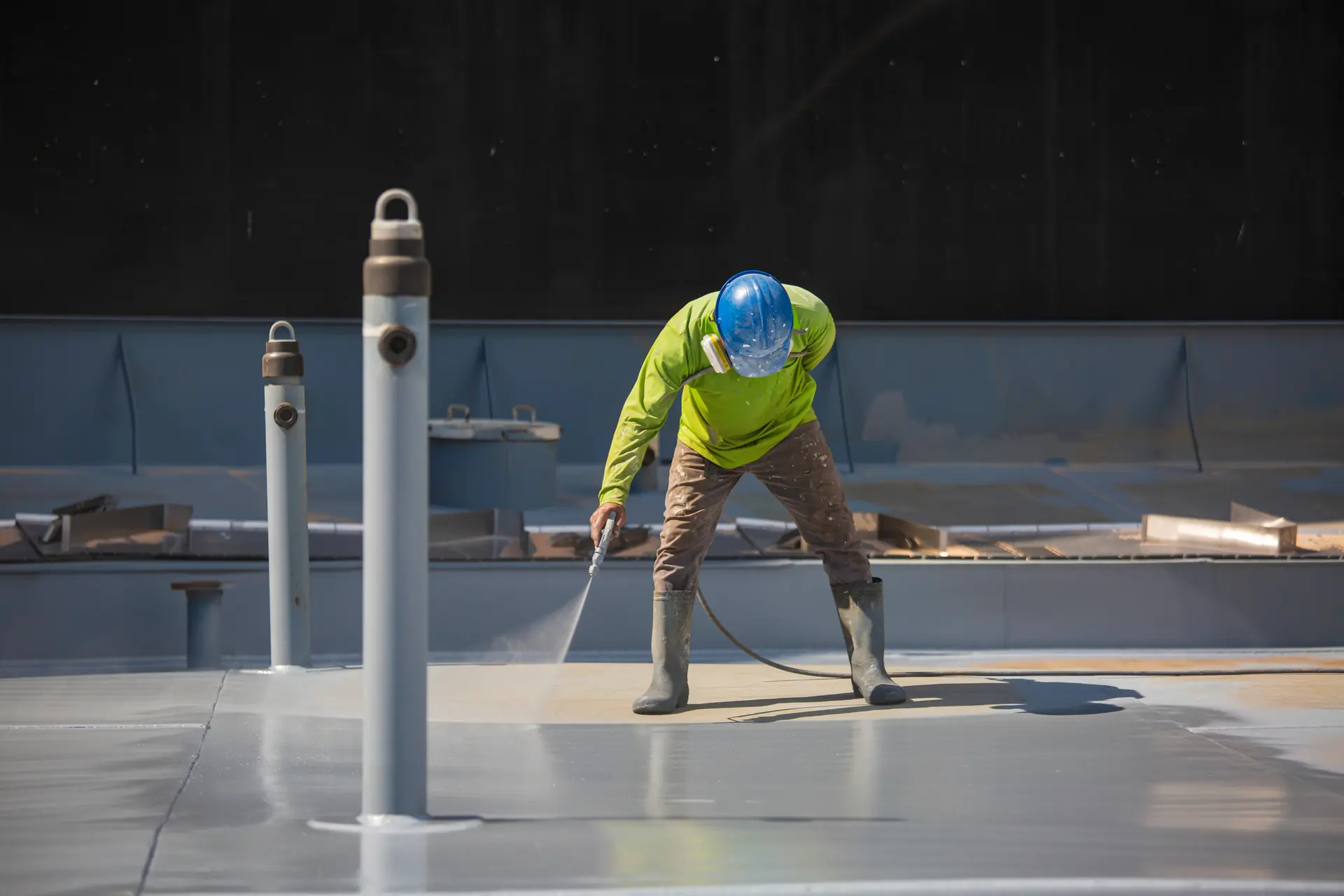 Construction worker in a blue helmet and green shirt spraying water or coating on a flat surface.