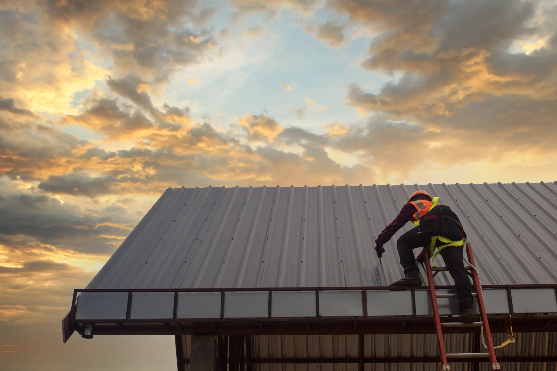 Worker in safety gear climbing a ladder to work on a metal roof at sunset with cloudy sky.
