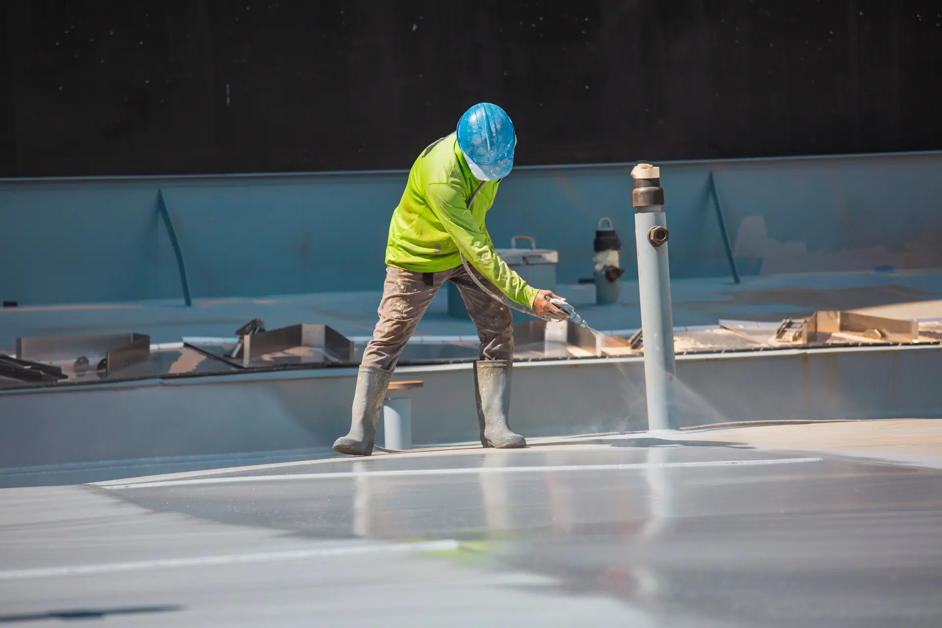 Construction worker wearing a green jacket, blue helmet, and rubber boots spraying water or liquid on a large flat surface.