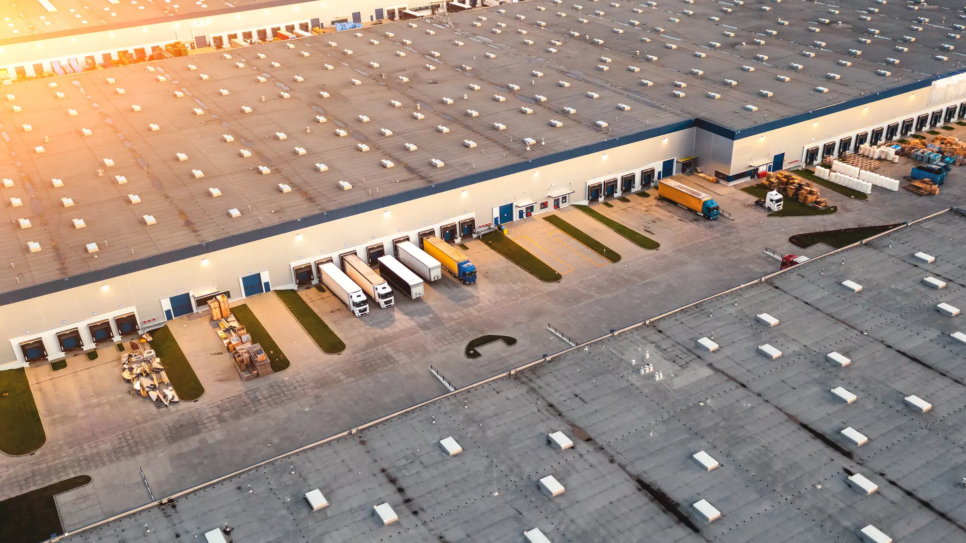 Aerial view of a large warehouse with several trucks backed up at loading docks during sunset.
