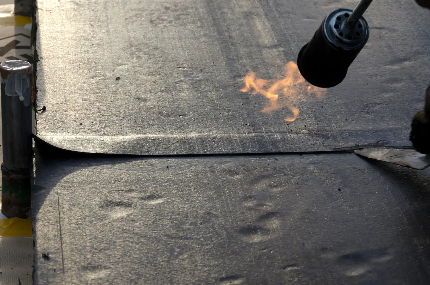 Worker using a blowtorch to heat and seal a black roofing membrane.
