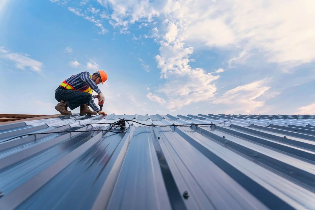 Construction worker in safety vest and helmet installing metal roofing panels under a partly cloudy blue sky.