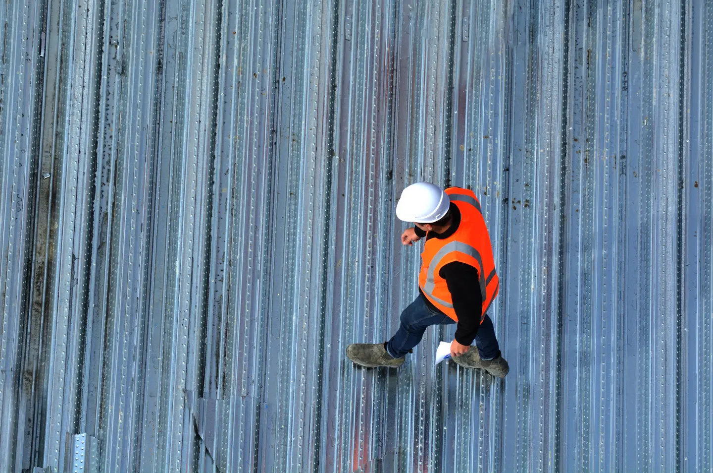 Construction worker in an orange safety vest and white hard hat walking on metal beams.