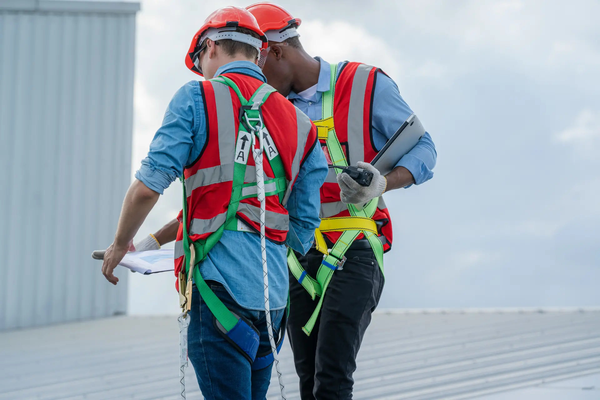 Two construction workers wearing safety harnesses and helmets reviewing documents on a metal rooftop.