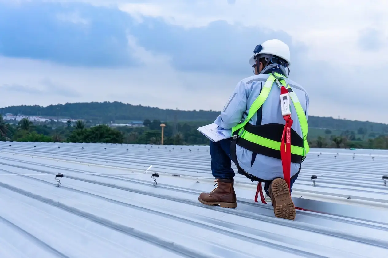 Construction worker wearing safety harness and helmet kneeling on a metal roof inspecting documents.