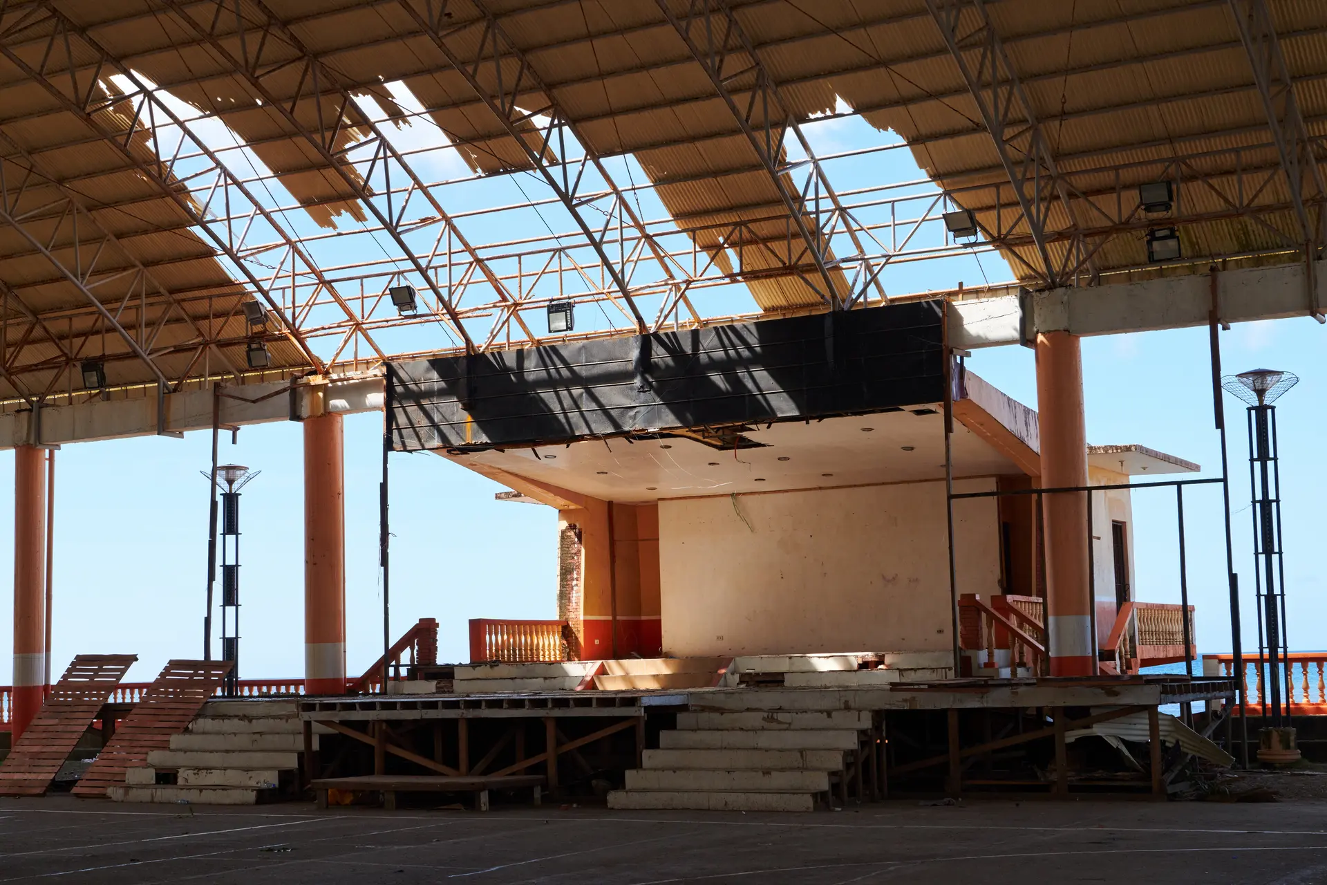 Abandoned indoor stage with damaged roof panels and worn stairs under a large metal-framed canopy.