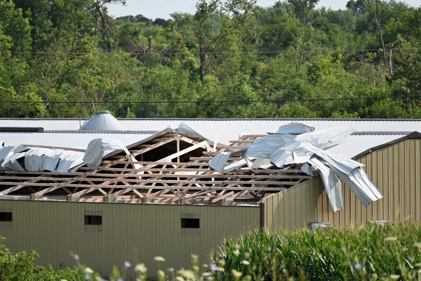 A metal-roof building with a partially destroyed roof, showing wooden beams and torn metal sheets, with trees in the background.