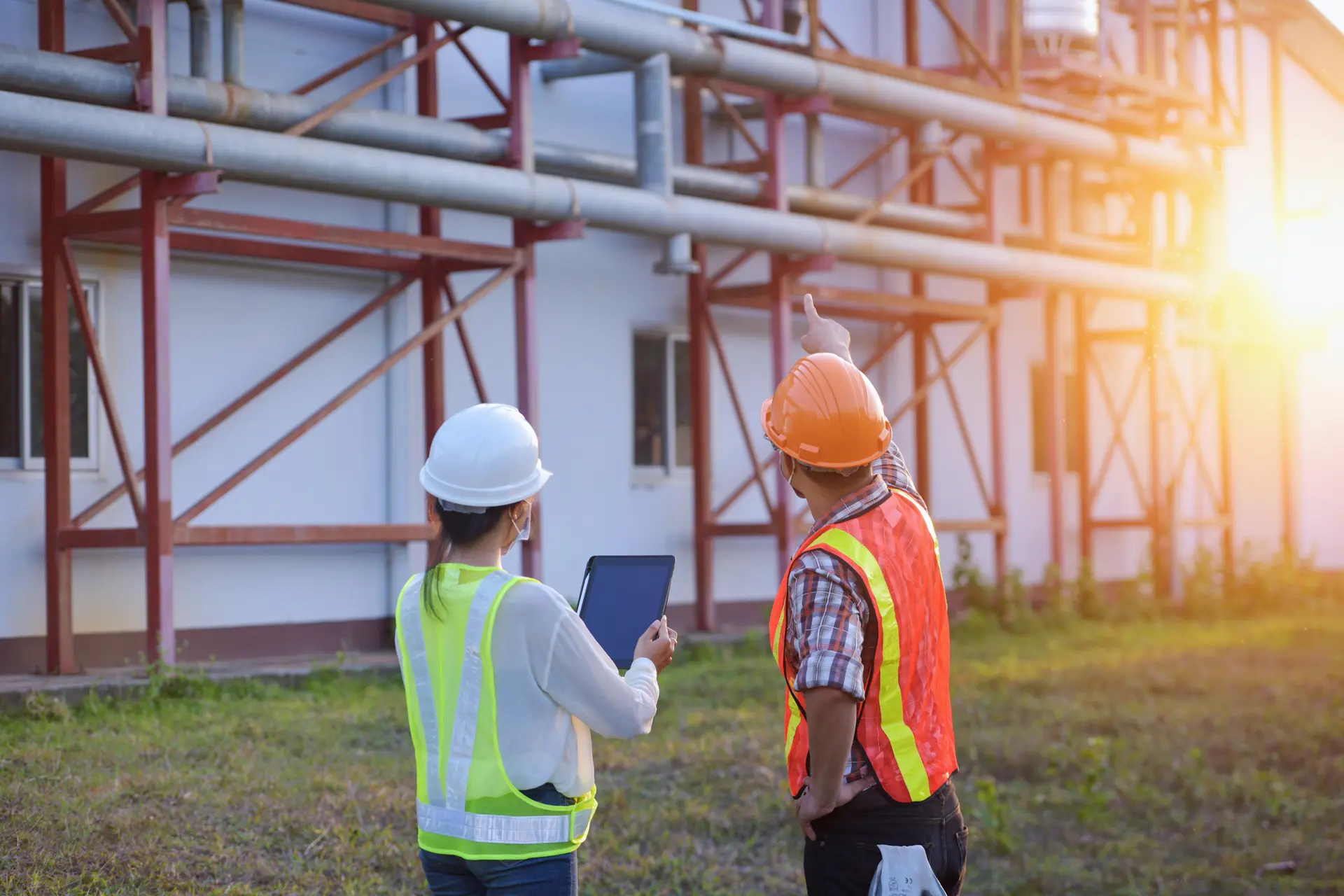 Two construction workers wearing hard hats and safety vests inspecting industrial pipes with one pointing and the other holding a tablet.