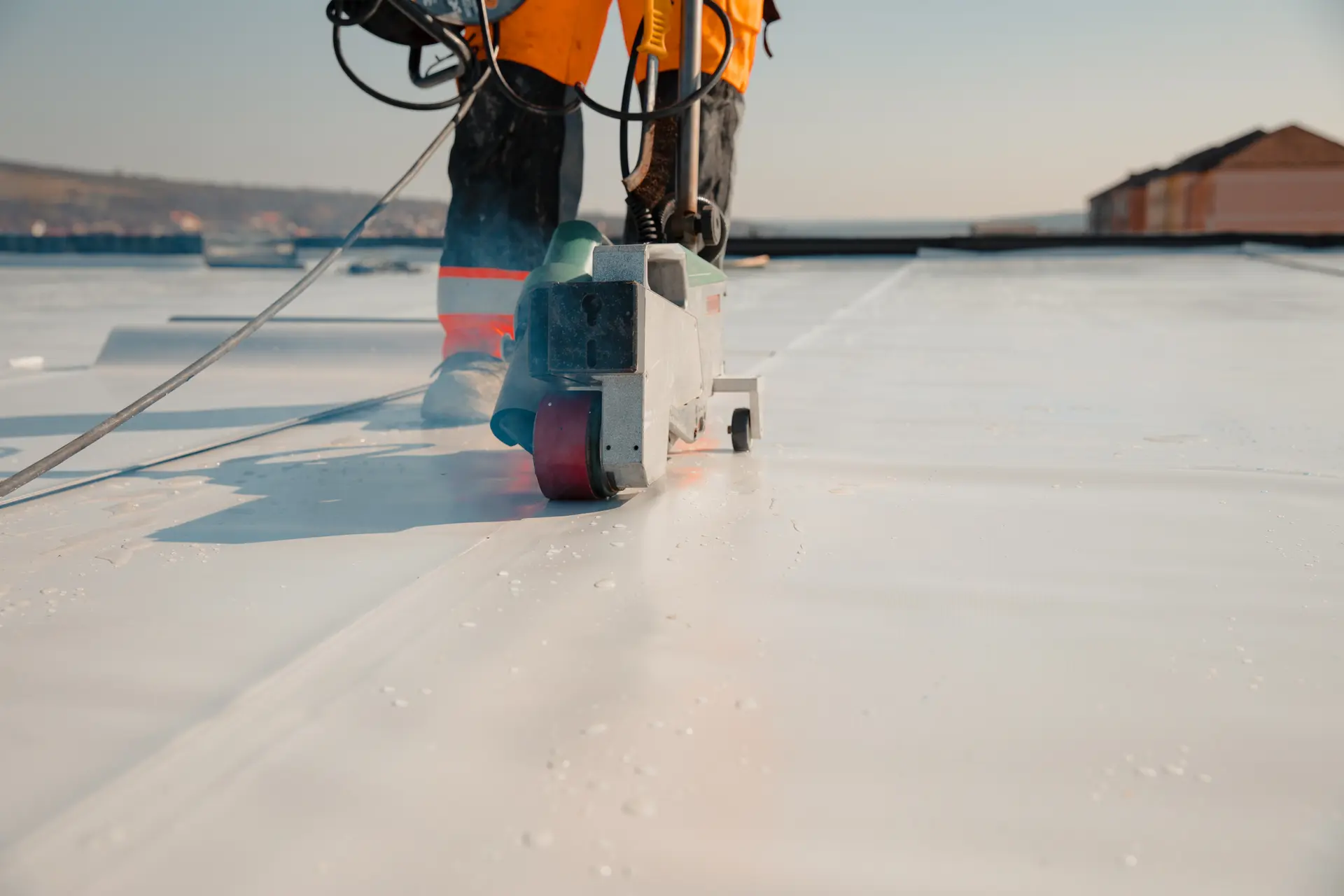 Worker in orange safety gear using a heat welding machine to seal white roofing membrane on a flat rooftop.