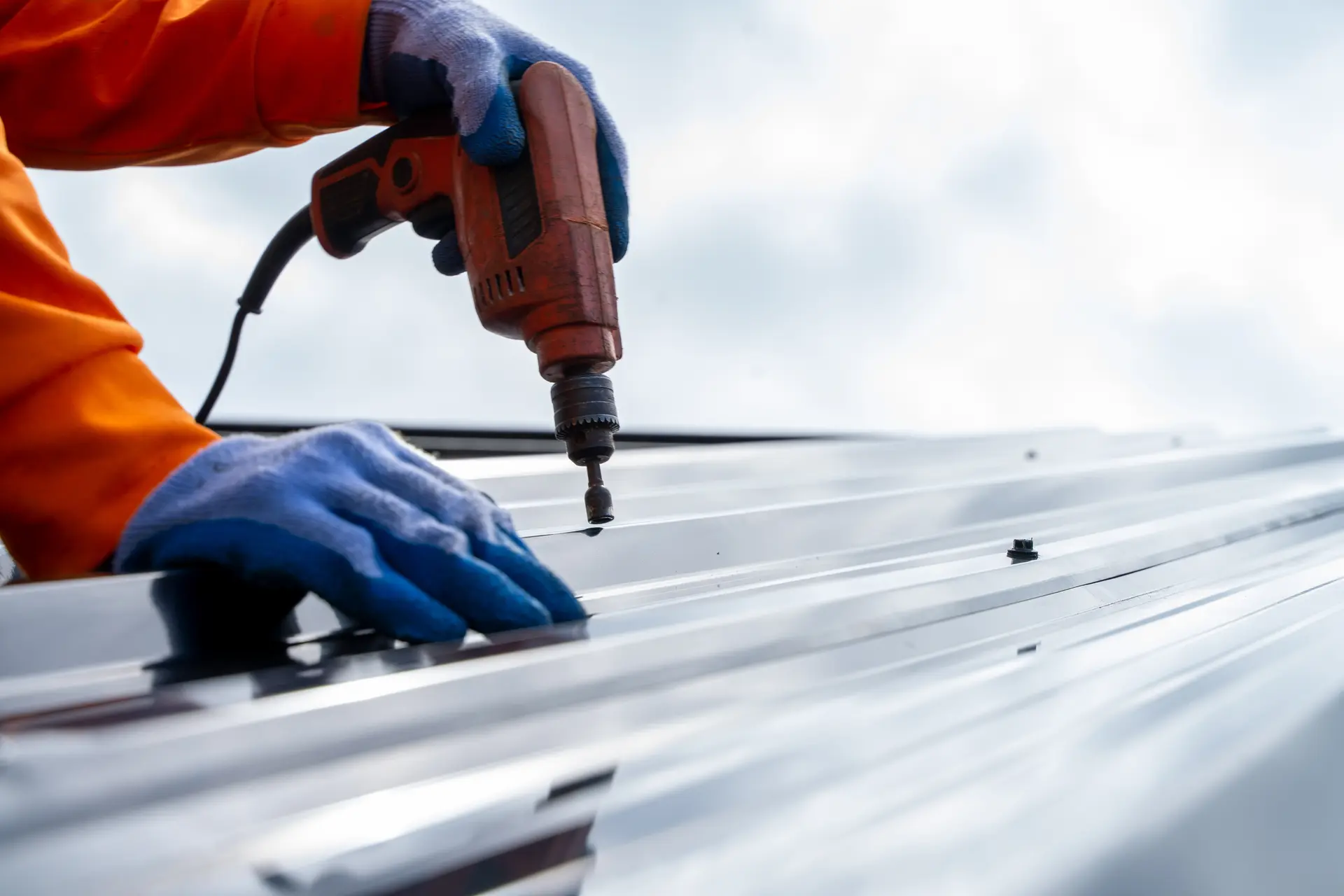 Person wearing blue gloves and orange jacket using a power drill on a shiny metal roof panel.