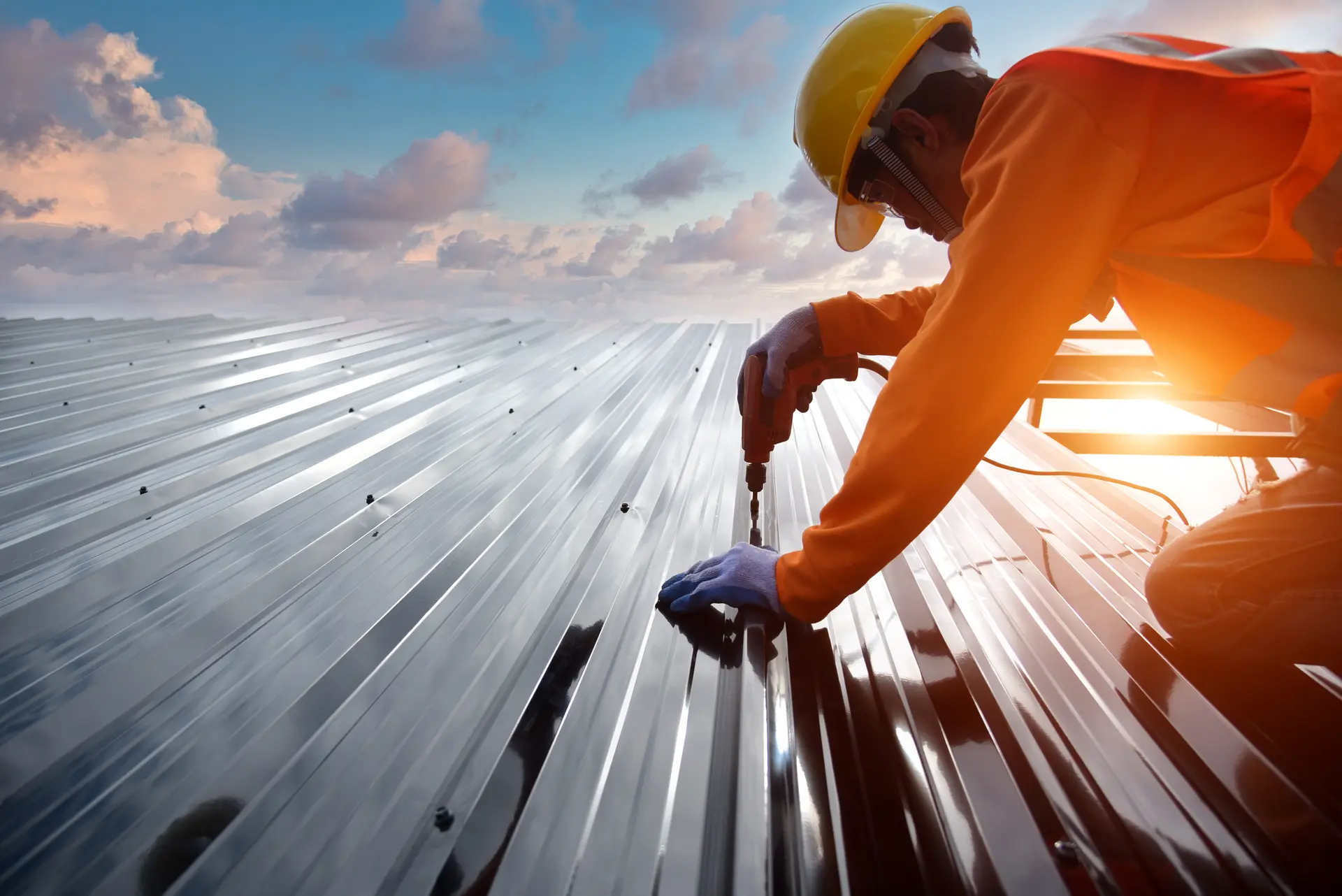 Construction worker in safety gear installing metal roofing panels with a power drill at sunset.