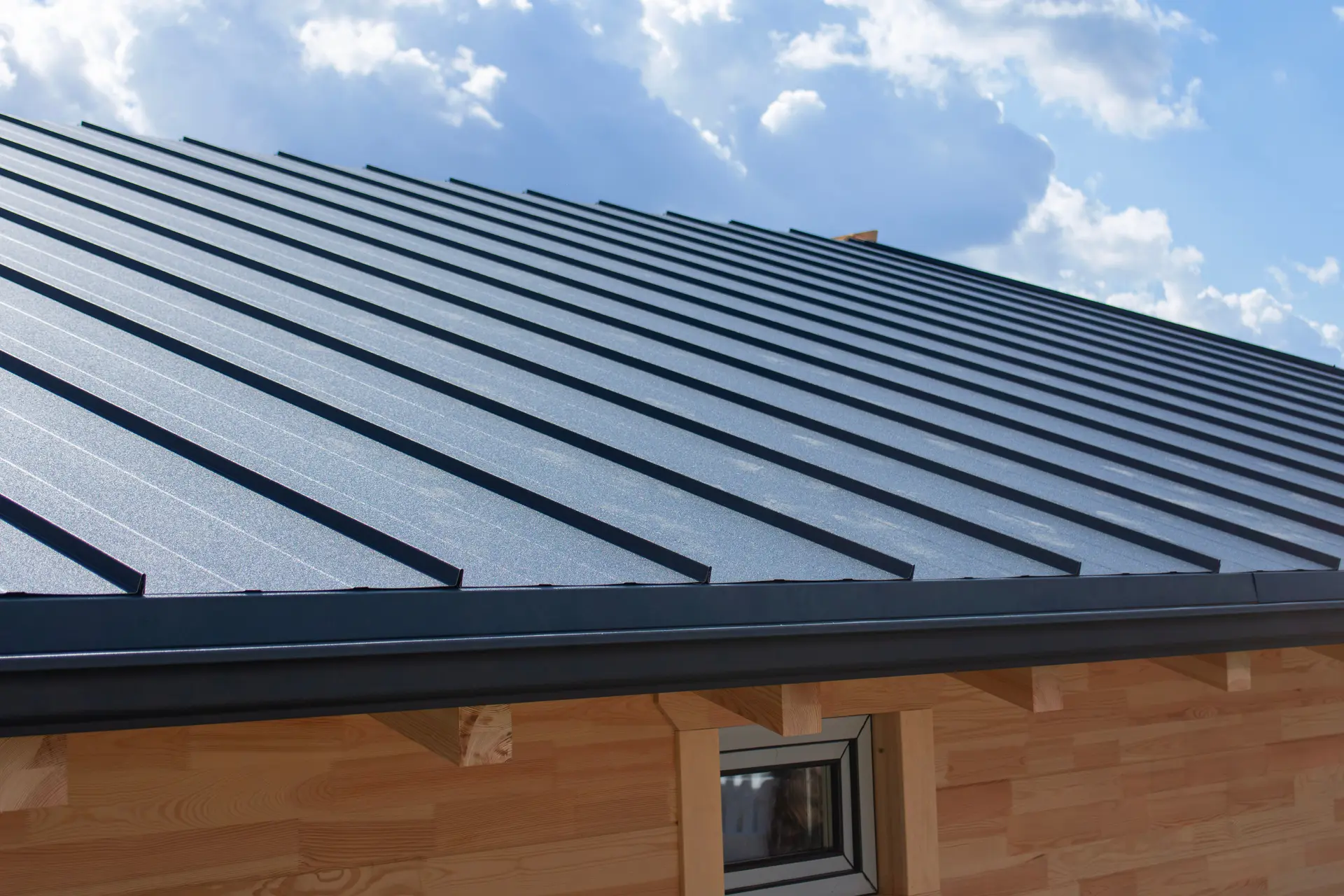 Close-up of a modern metal roof with dark panels and wooden wall under a partly cloudy sky.