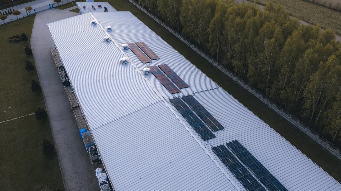 A large industrial building roof with several solar panel arrays and ventilation units, bordered by trees and a fenced area.